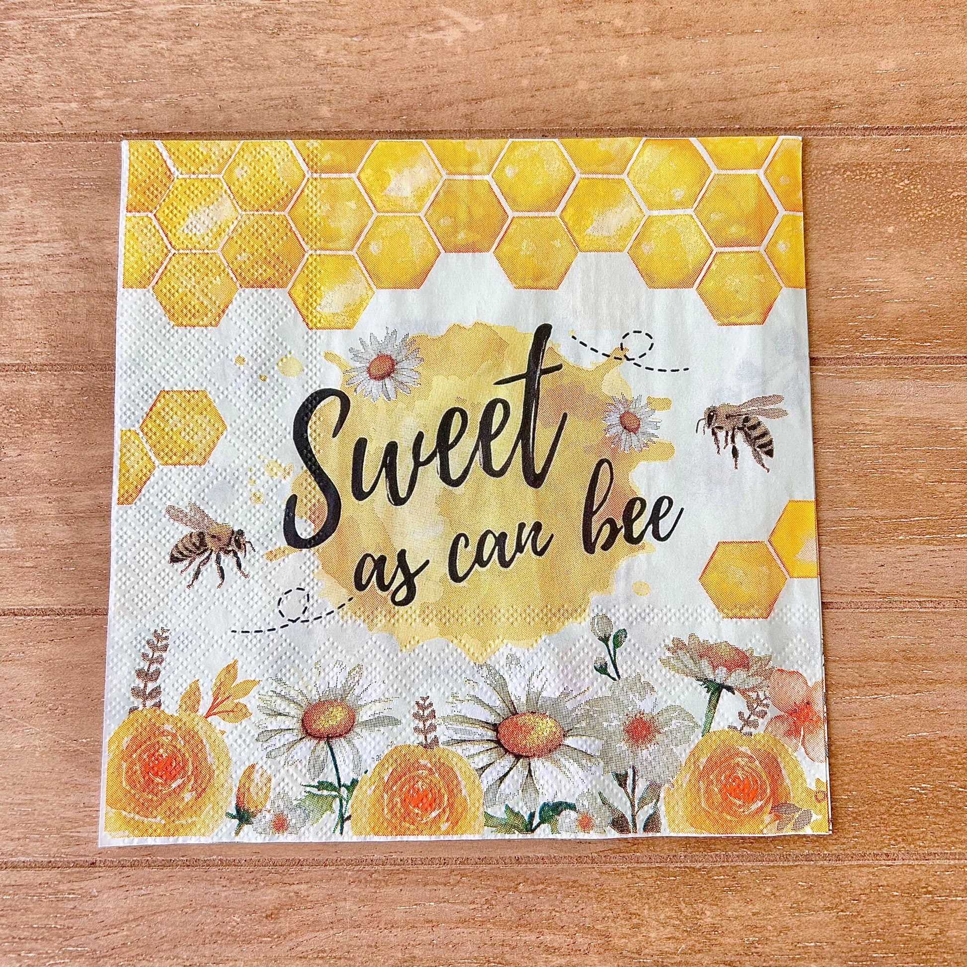 Napkin with honeycomb design, flowers, and text 'Sweet as can bee' on a wooden surface