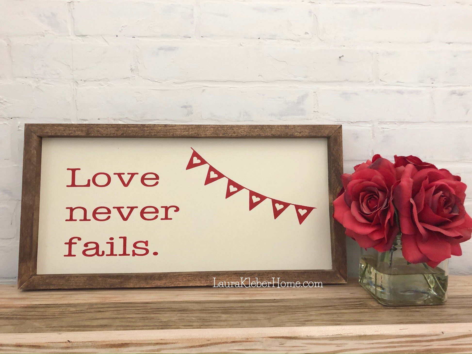 A 10x18 inch framed wood sign with love never fails painted in red with a white background.
