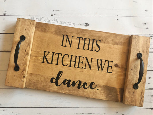 A 10x18 inch wood tray completed in Early American stain with the words In this kitchen we dance in black with black metal handles.